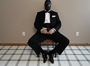 Seated in a tuxedo wearing a rubber cock and ball